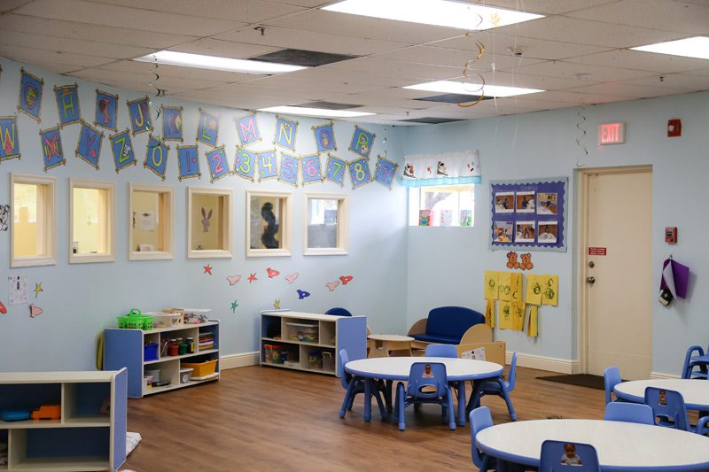 Brightly colored preschool classroom featuring learning materials, tables, and decorative alphabet banners on the walls.